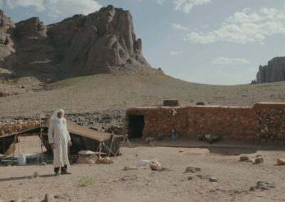 A man standing beside a tent and stone shelter in an arid mountain landscape.
